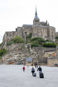 people walking with luggage towards a large abbey on a hilltop