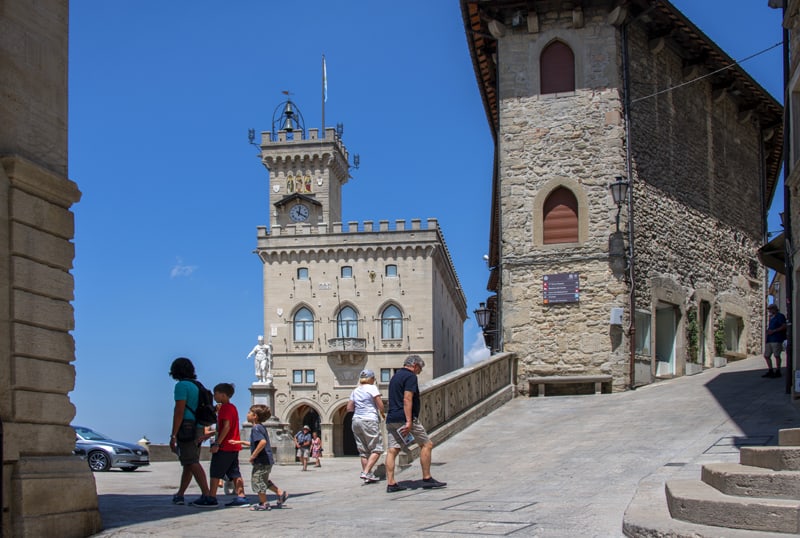 people walking by old stone buildings on a plaza