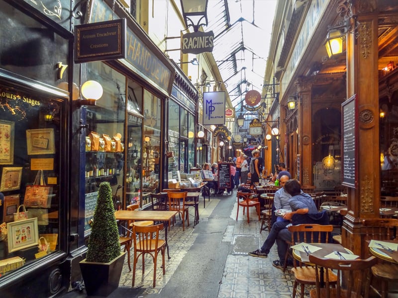 people in a long narrow hallway sitting at tables in one of the hidden passages of Paris