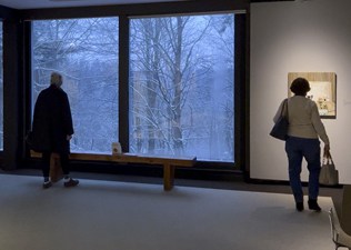 a woman looking at a painting on the wall as another woman looks out a large window at a snow-covered forest