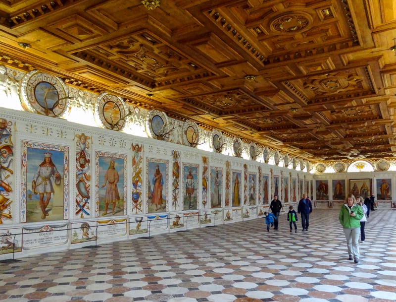 people walking through a large hall with many paintings and a carved wooden ceiling
