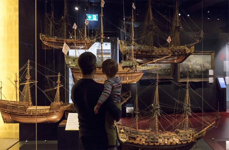 man holding a child looking at wooden ship models