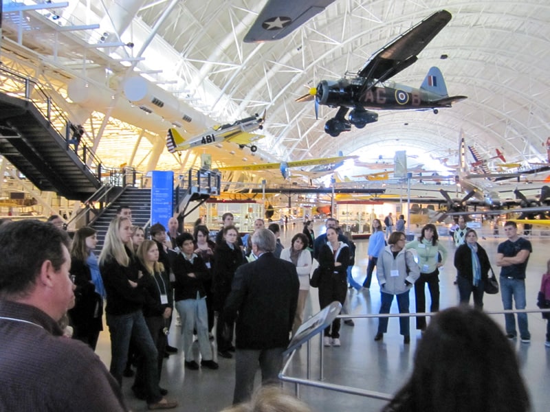 A group of people listening to a museum docent by a large group of airplanes in the Air Space Museum Dulles
