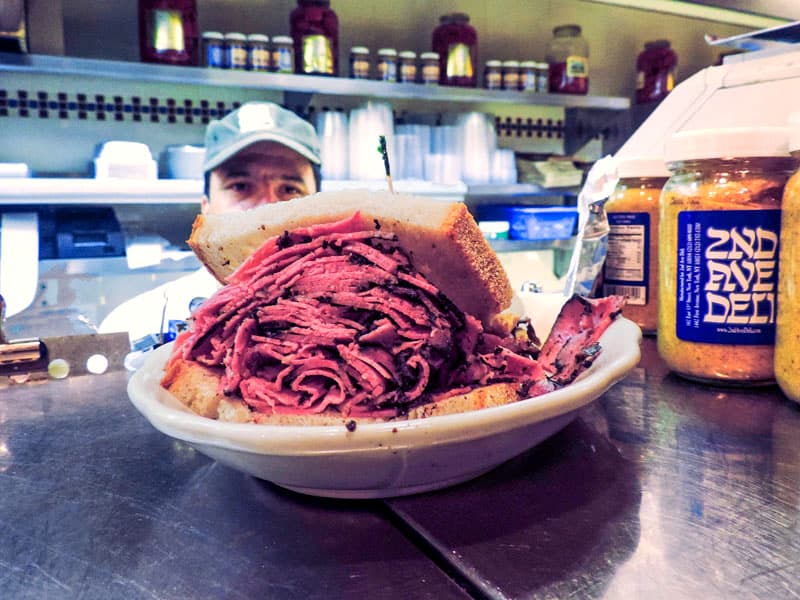 a chef in a kitchen looking at a just-made pastrami sandwich