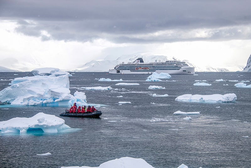 people in red jackets on a raft moving through sea ice with a large ship in the background