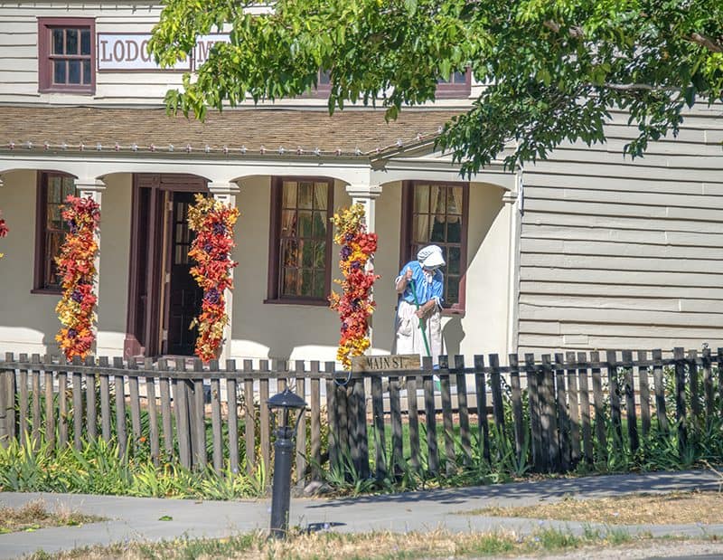 a woman in a bright blue dress sweeping the porch of a lodge in this is the place heritage park, one of the things to do in salt lake city