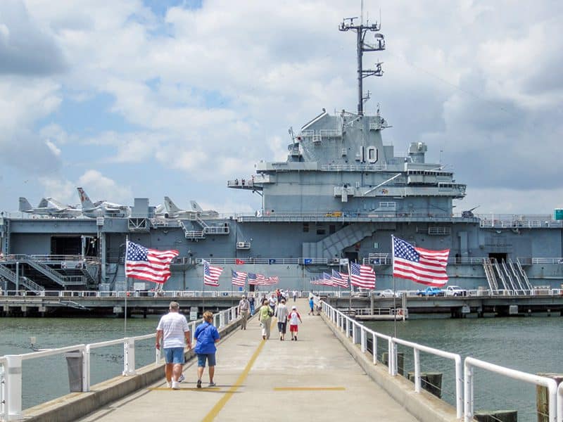 people walking on a pier towards a museum ship with American flags flying
