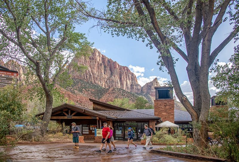 people walking past a visitor's center in a Utah National Parks with a tall mountain in the distance