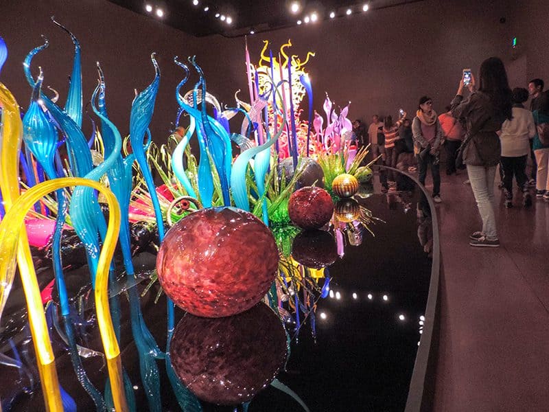 people walking past an exhibit of brightly colored glass