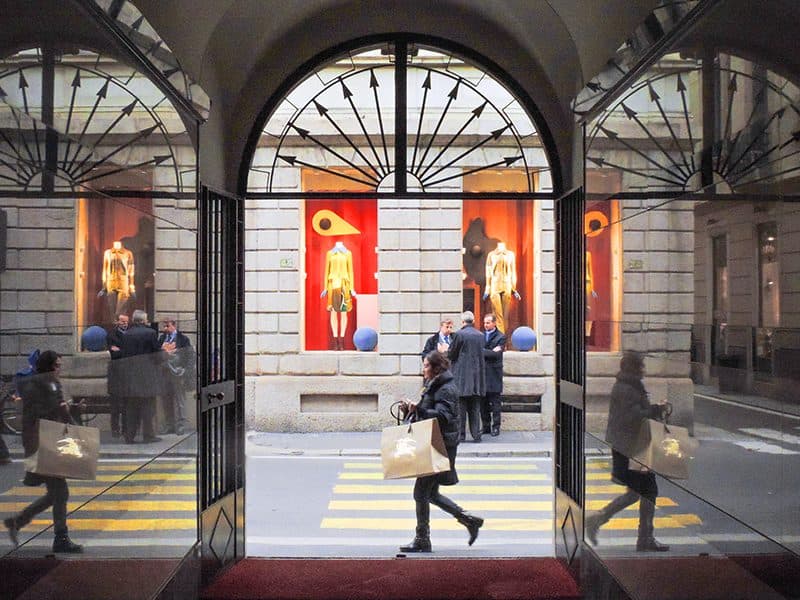 a woman walking past ornate shop windows by which a group mof men are standing and talking