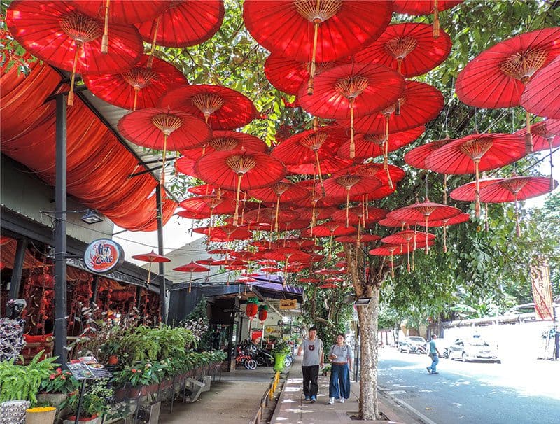 a couple walking along a sidewalk beneath red umbrellas hanging from a tree