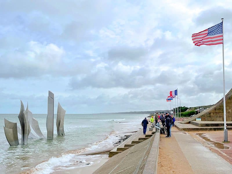 people standing beneath an American flag looking at a modern sculpture off a beach